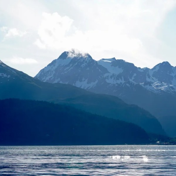 Mountain range with snow and clouds above a calm lake under a bright sky.