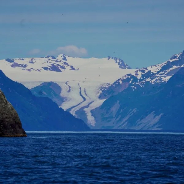 Snowy mountains and rock formations above a blue sea under a clear sky.