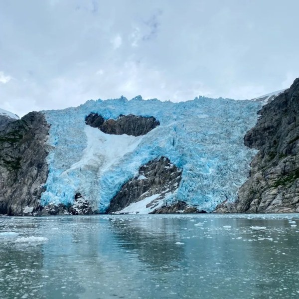 Glacier with blue ice between rocky cliffs, viewed across calm, icy water.