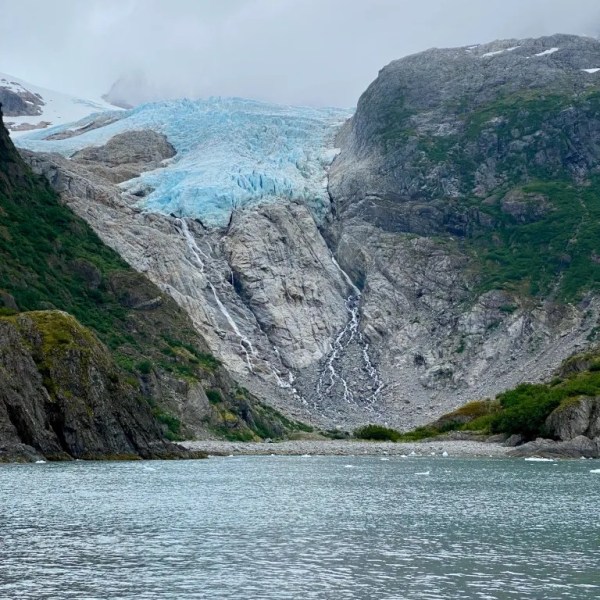 Blue glacier between rocky mountains with water in foreground.