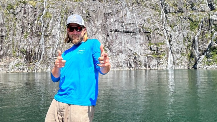 Person on a boat in front of rocky cliffs and water, pointing and smiling in sunny weather.