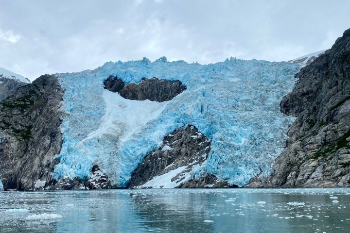 Large blue glacier between rocky cliffs over calm water under cloudy sky.