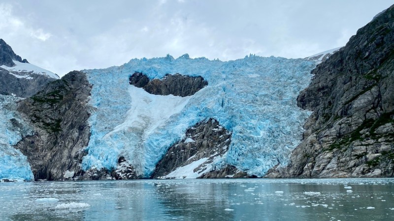 Large blue glacier between rocky cliffs over calm water under cloudy sky.