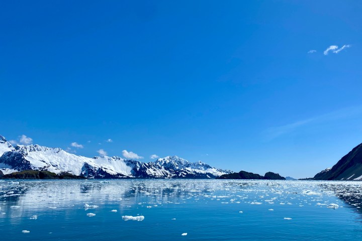 Snowy mountains and icebergs reflecting on a calm blue sea under a clear blue sky.