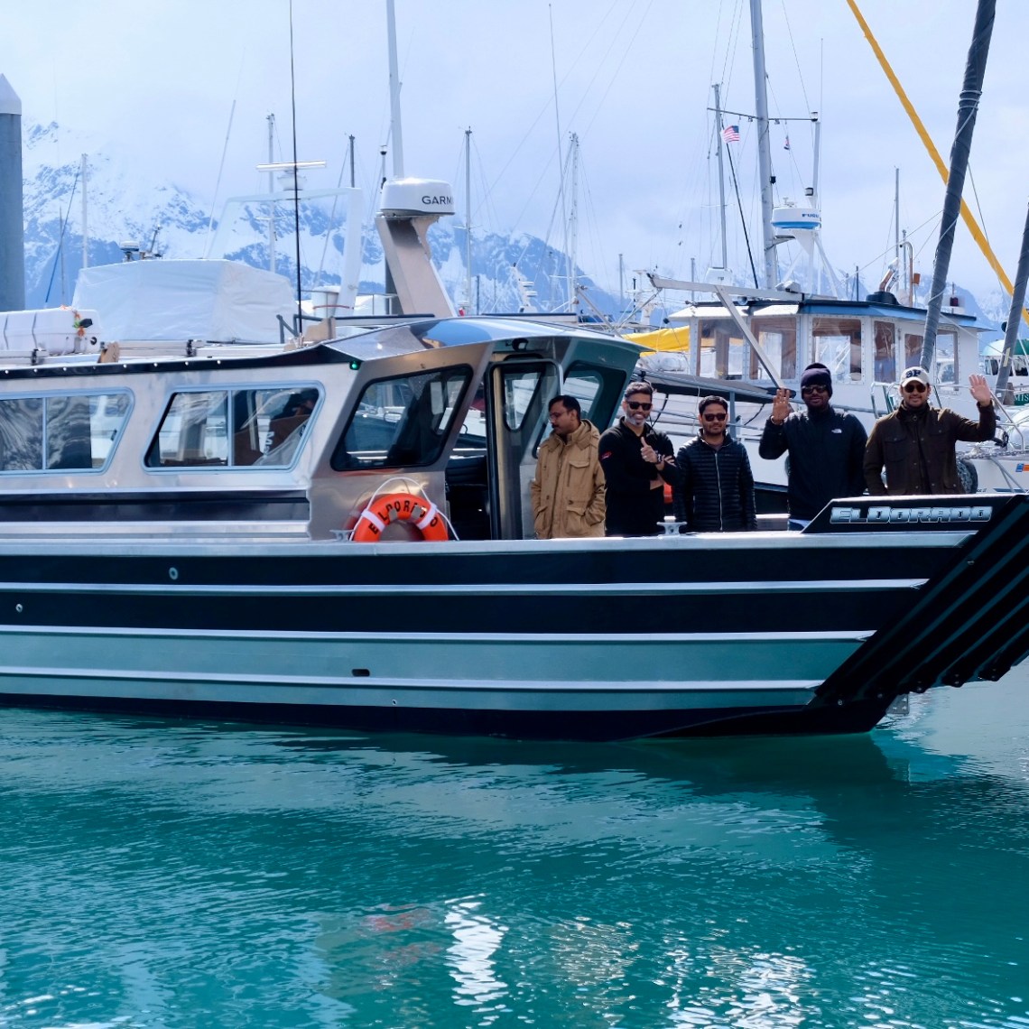 Group of people on a docked boat in a marina with mountains in the background.